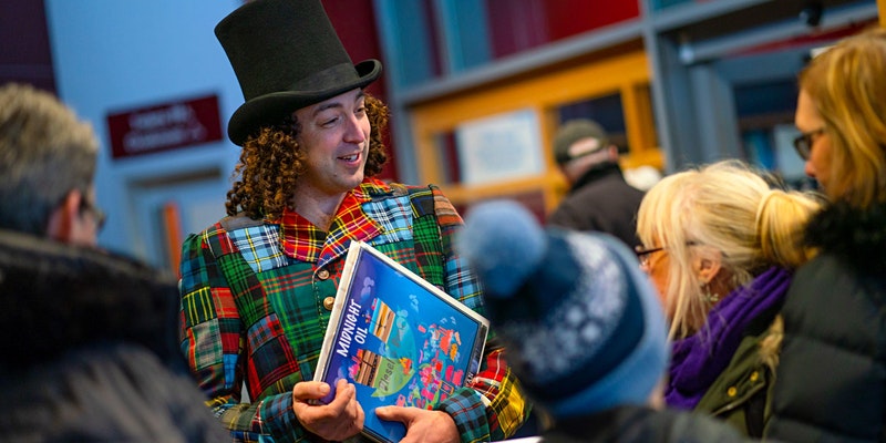 A photo of the Rock Showman, from the waist up holding a blue children's book titled 'Midnight Oil'. He wears a black top hat and multi colour tartan tails. He is stood talking with members of the public - adults and children, with their heads facing away from the camera in the foreground.