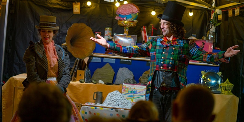 A photo of the Rock Showman and Floss facing the crowd. The Rock Showman wears a top hat and multicolour tartan tails. Floss wears a pink scarf, a grey ruffled blazer and beige hat. In the background is their set, which features various rocks, minerals and ores, plus an old grammarphone.