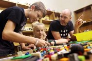 A photo of a man and two children sat at a table, as the children play with LEGO pieces on the table.