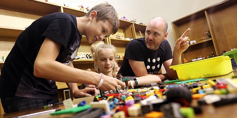 A photo of a man and two children sat at a table, as the children play with LEGO pieces on the table.