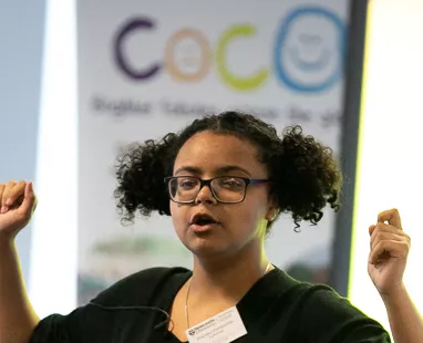 A photograph of a woman talking, with her hands raised. She stands in front of a banner with COCO's logo on.