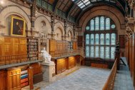 A photograph of The Common Room's Wood Hall featuring colourful stained glass windows, wooden bookcases along the walls and balconies (some are open, so you can see some of the books), a large portrait hanging from the wall, and a large white statue of a man in the centre of the room.