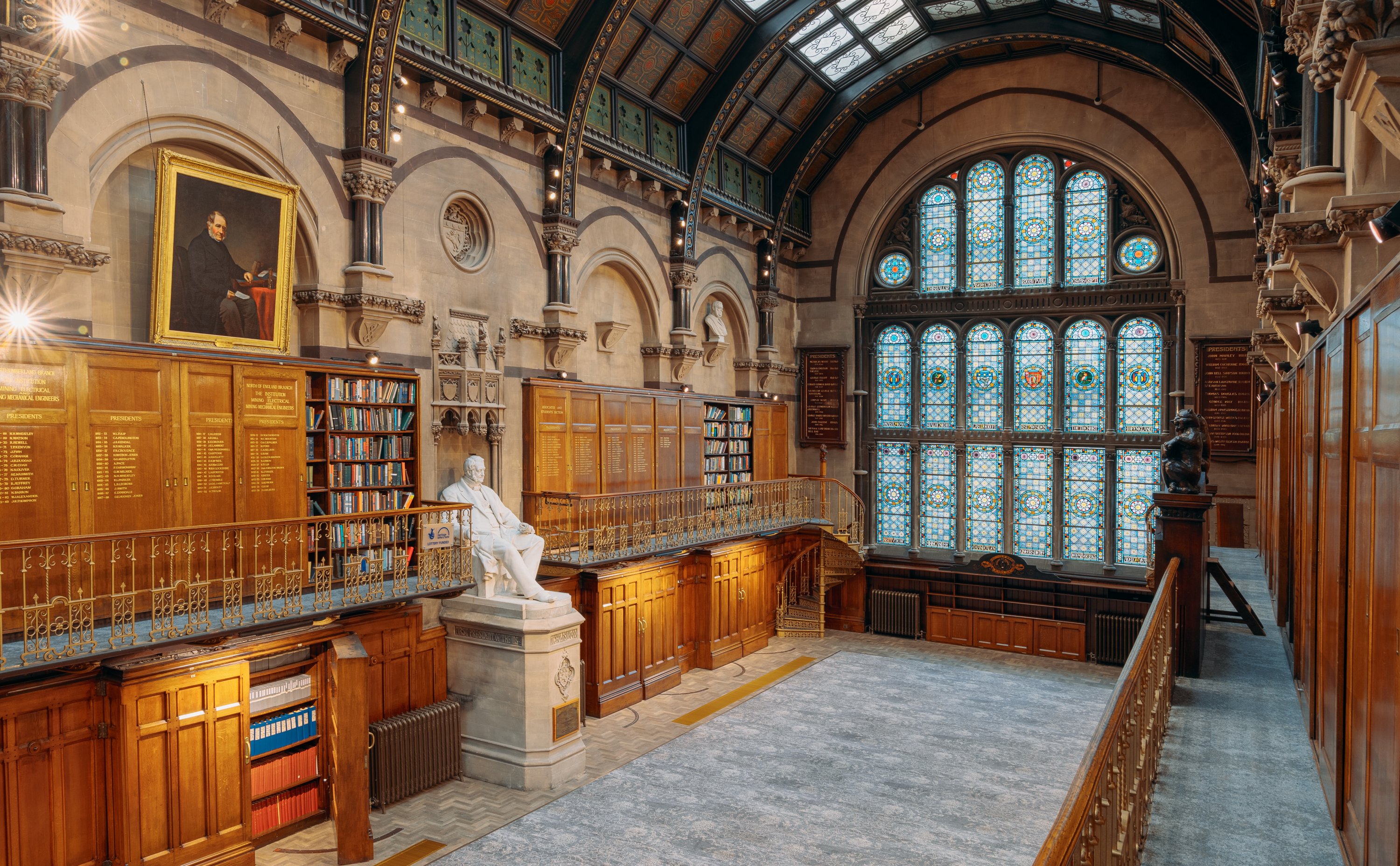 A photograph of The Common Room's Wood Hall featuring colourful stained glass windows, wooden bookcases along the walls and balconies (some are open, so you can see some of the books), a large portrait hanging from the wall, and a large white statue of a man in the centre of the room.