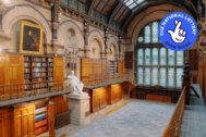 A photo taken in The Common Room's Wood Hall. A grand hall line with wooden bookcases. Balconies run along either side of the hall, with golden railings. At the end of one balcony, you can see a golden spiral staircase. In the middle of the room is a large white statue on a plinth of Nicholas Wood. An arched stained glass window is at the end of the room. The room has a curved, glass ceiling, and a large portrait is seen hanging above one of the bookcases. The National Lottery Open Week logo sits on top of the image, in the top-right corner.