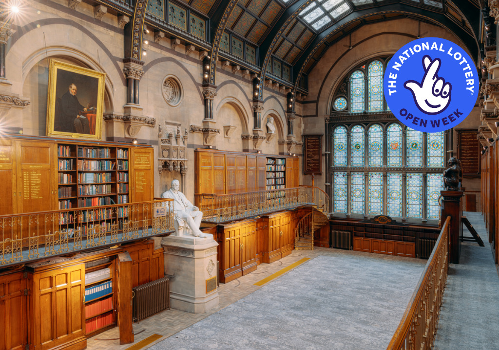 A photo taken in The Common Room's Wood Hall. A grand hall line with wooden bookcases. Balconies run along either side of the hall, with golden railings. At the end of one balcony, you can see a golden spiral staircase. In the middle of the room is a large white statue on a plinth of Nicholas Wood. An arched stained glass window is at the end of the room. The room has a curved, glass ceiling, and a large portrait is seen hanging above one of the bookcases. The National Lottery Open Week logo sits on top of the image, in the top-right corner.